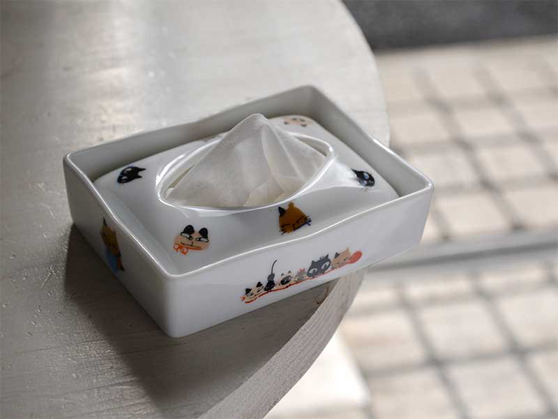シンジカトウさんがデザインした可愛い猫のイラストが白い陶磁器のポケットティッシュケースに描かれ、白いテーブルの上に置いてある画像です。