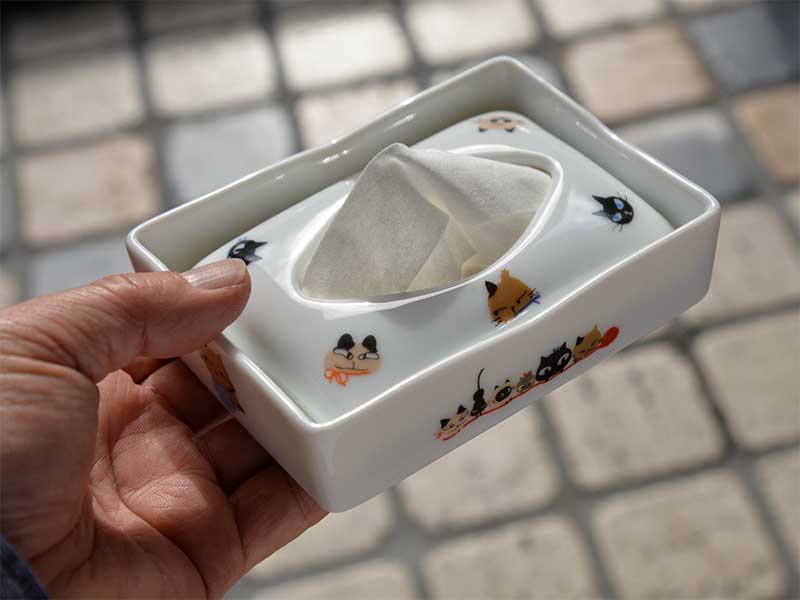 シンジカトウさんがデザインした可愛い猫のイラストが白い陶磁器のポケットティッシュケースに描かれ手にもって大きさを比較できる画像です