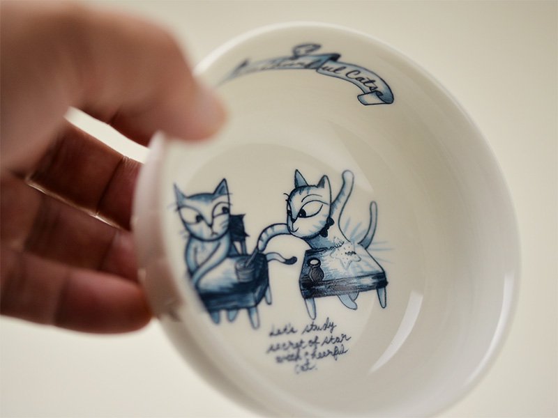 雑貨デザイナーシンジカトウさんが勉強するイタズラ好きな猫達のイラストを直径10cm位の陶磁器製のボウルに描いたボウル、小鉢を手に取った様子の画像です。