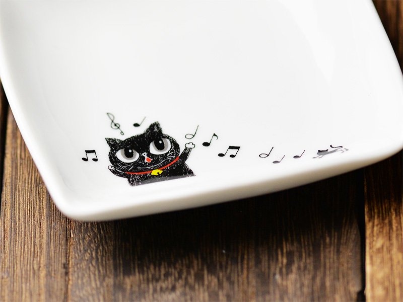 美濃焼製の白い小皿に雑貨デザイナーシンジカトウさんが黒猫が楽しそうに歌う様子のイラストを描いた小皿のをデザインが分かるようにデザイン部分を大きく拡大した画像です。