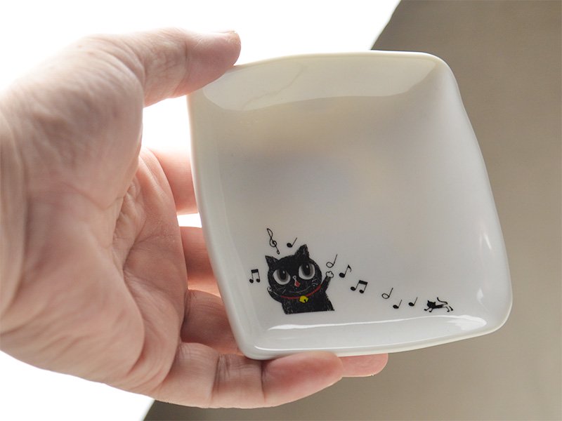 美濃焼製の白い小皿に雑貨デザイナーシンジカトウさんが黒猫が楽しそうに歌う様子のイラストを描いた小皿を手に取った時の様子の画像です。