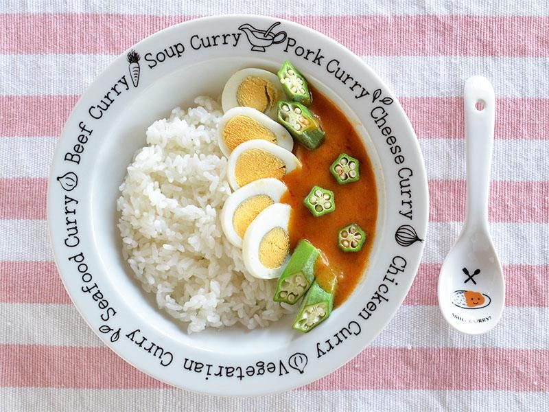 「スマイルカレーシリーズ カレー&パスタプレート」に盛られた色鮮やかなカレーライス。左半分には白いご飯、右半分には卵とオクラがトッピングされたオレンジ色のカレーが盛り付けられており、食欲をそそる一皿です。プレートのリムには様々な種類のカレー名と可愛いイラストが描かれています。雑貨デザイナーシンジカトウ氏による、かわいいデザインです。