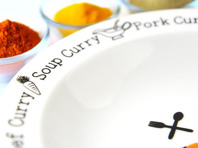 スマイルカレーシリーズ カレー&パスタプレート」の白いリム部分のクローズアップ画像。リムには「Soup Curry」の文字と可愛らしいカレーポット、そして「Carrot Curry」の文字と人参のイラストが黒字でスッキリと描かれており、背景には鮮やかなカレー粉が写っています。雑貨デザイナーシンジカトウ氏の可愛いイラストがポイントです。