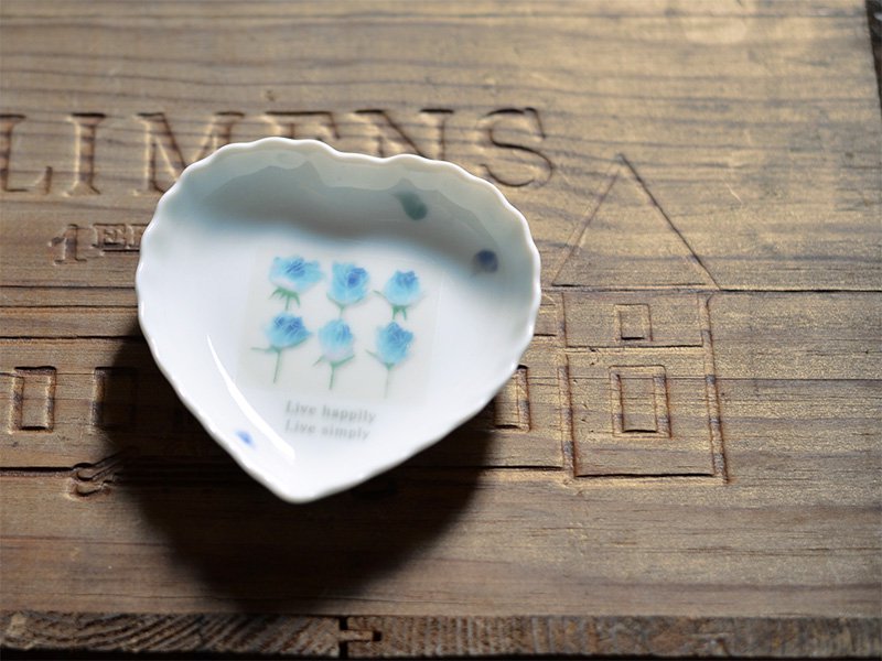 シンジカトウさんがデザインしたブルーローズのイラストが描かれた白い陶磁器製のハートの形をした小皿をアンティークなボードに乗せた様子を撮影した画像です。