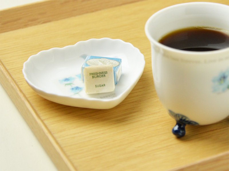 シンジカトウさんがデザインしたブルーローズのイラストが描かれた白い陶磁器製のハートの形をした小皿に角砂糖をのせてコーヒーカップと一緒に撮影した画像です。