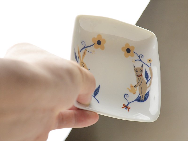 美濃焼製の白い小皿に雑貨デザイナーシンジカトウさんが猫とうさぎのイラストを描いた小皿を手に取った時の様子の画像です。