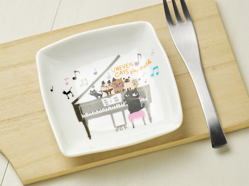 美濃焼製の白い小皿に雑貨デザイナーシンジカトウさんが黒猫がピアノを弾き5匹の猫が鍵盤の上で合唱するユーモラスなイラストを描いた小皿の画像です。