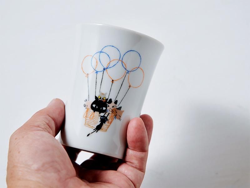左手でタンブラーを優しく持っている写真。不揃いな猫たちがバスケットに乗って風船で空を飛んでいるイラストが大きく写っており、デザインの細部や陶器の質感がよくわかります。